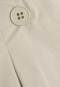 Béžová látka s hladkým povrchem, která má velký kulatý knoflík se čtyřmi dírkami, přišitý na levé straně.
