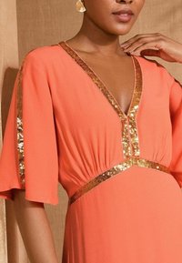 Koraalroze jurk met een V-halslijn, uitlopende mouwen en gouden sequins langs de halslijn en taille. Lichtgewicht stof, zachte textuur.