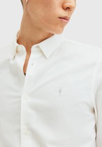 Hvid skjorte med knapper i et struktureret stof, med et lille gråt emblem på brystet og et klassisk kravedesign.