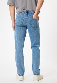 Jeans de mezclilla azul claro con diseño de pierna recta, costuras visibles y dos bolsillos traseros. Llevado con una camisa gris y zapatillas blancas.