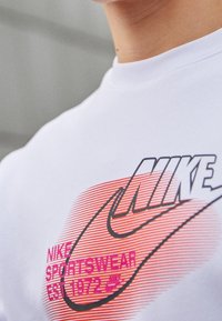 Vit Nike-tröja med en stor svart swoosh-logotyp, rosa och orange gradientränder, samt texten: "NIKE SPORTSWEAR EST. 1972."