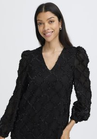 Svart texturerad klänning med V-ringning, puffiga långa ärmar och blommig applikerade detaljer. Har ett mönstrat design med upphöjda former.