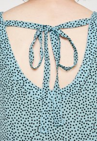 Top senza maniche di un azzurro chiaro con schiena scoperta, caratterizzato da pois neri e una chiusura a fiocco con due lunghe e fluide laccetti.
