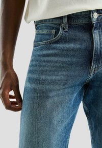 Dorosły w niebieskich dżinsach i białej koszuli, z ręką luźno opadającą wzdłuż ciała, pokazany od talii do połowy uda na jednolitym tle.