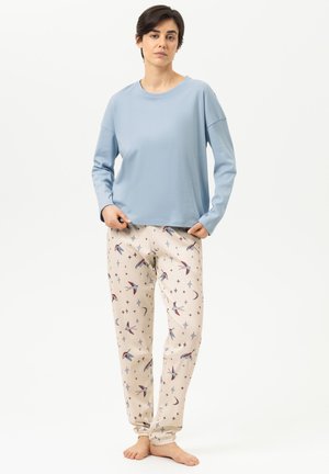 Vrouw staat blootsvoets, draagt een lichtblauw shirt met lange mouwen en crèmekleurige broek met vogel- en sterrenpatronen tegen een witte achtergrond.