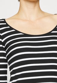 Person trägt ein schwarz-weiß gestreiftes Oberteil mit horizontalen Streifen und einem weiten, abgerundeten Ausschnitt, der den Hals und den oberen Brustbereich zeigt.