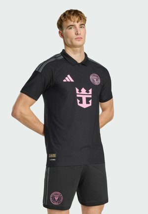 Junger Mann, der ein schwarzes Trikot und Shorts des Club Internacional de Fútbol Miami mit pinkfarbenen Logos trägt, steht mit den Händen hinter dem Rücken.