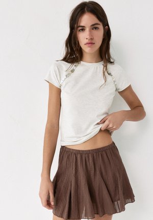 Mladá žena s dlouhými hnědými vlasy, oblečená v bílém tričku s krátkými rukávy se flitry a hnědé plisované sukni, stojící před jednoduchou bílou stěnou.