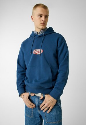 Giovane uomo che indossa una felpa con cappuccio blu con logo rosso, sopra una camicia con colletto, in piedi mentre si aggiusta la cintura dei jeans con le mani.