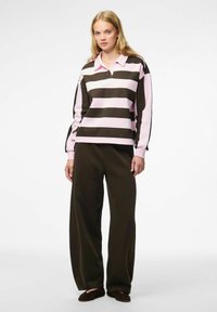 Femme debout portant un polo à manches longues rayé rose et marron, un pantalon ample marron foncé et des chaussures marron foncé sur fond blanc.