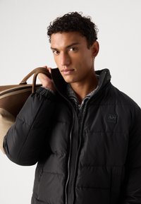 Chaqueta acolchada negra con cremallera, cuello alto y un discreto parche de logo. Lleva puesta sobre un suéter texturizado gris, complementado con un bolso de hombro beige.