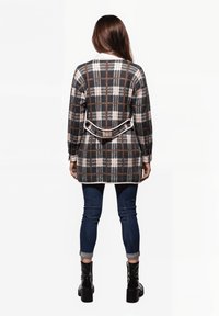Larga chaqueta de cuadros en gris oscuro, blanco y marrón, con un detalle de cinturón en la cintura, mangas ajustadas y combinada con jeans azules y botas negras.