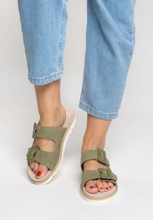 Füße in hellgrünen Sandalen mit Schnalle, weißen Sohlen und rotem Nagellack, kombiniert mit hellblauen, gekürzten Jeans.