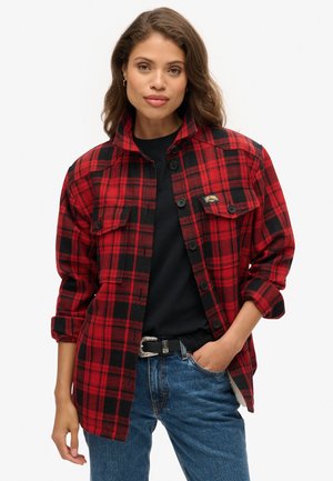 Chemise en flanelle à carreaux rouges et noirs, style à boutons avec des manches longues, deux poches sur la poitrine, portée sur un haut noir et un jean bleu.