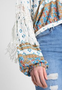 Bluzka w stylu boho z długimi białymi frędzlami, zdobiona wielokolorowym kwiatowym i geometrycznym wzorem na jasnym tle; marszczone rękawy.