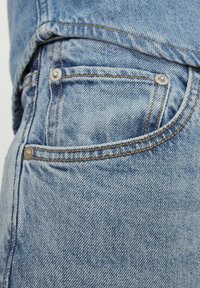 Közelkép világoskék farmernadrágról, amelyen a hátsó zseb látható fém szegecsekkel és narancssárga varrással.