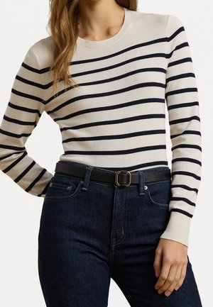 Femme portant un pull beige à manches longues avec des rayures horizontales noires, un jean bleu foncé et une ceinture noire avec une boucle dorée.