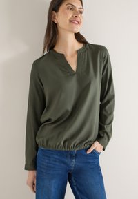 Blouse vert olive avec encolure en V et ourlet froncé, en tissu lisse. Manches longues et coupe décontractée, associée à un jean bleu.