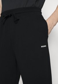 Pantalones jogger negros con cintura ajustable, bolsillos laterales y logo blanco "HUGO" en el muslo, fabricados con un tejido suave y ligero.