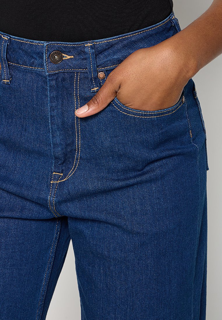 Jean en denim bleu foncé avec une taille haute, des accents de couture visibles et une poche sur le côté droit. Le tissu semble robuste avec une texture lisse.