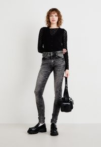 Un haut noir en dentelle à manches longues sous un gilet noir, associé à un jean gris taille haute et des chaussures noires imposantes. Un petit sac noir est tenu dans une main.