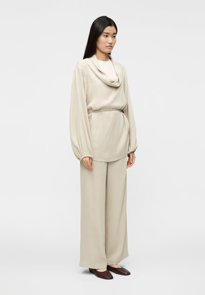 Μπεζ φαρδιά μπλούζα με ντραπέ λαιμόκοψη και μακριά μανί κια, σε συνδυασμό με φαρδιά ψηλόμεσα παντελόνια και σκούρα καφέ φλατ παπούτσια.