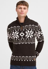 Brauner Strickpullover mit hohem Kragen, verziert mit weißen und grauen Schneeflockenmuster und einem braunen Reißverschlussakzent. Gerippte Bündchen und Saum.
