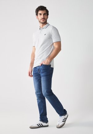 Polo a maniche corte grigio chiaro con dettagli neri, abbinato a jeans blu scuro e sneakers bianche con strisce nere e dettagli grigi.