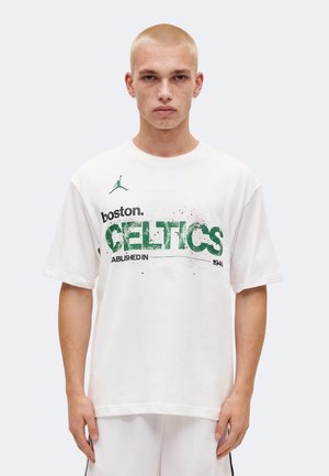 Joven con cabello rubio corto que lleva una camiseta blanca de Boston Celtics y pantalones cortos blancos, de pie contra un fondo liso y claro.