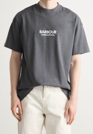 Osoba ubrana w ciemnoszarą koszulkę Barbour International i jasnobeżowe spodnie, stojąca na jednolitym białym tle.