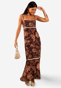Mujer sonriendo, vestida con un maxi vestido floral marrón de tirantes finos, sosteniendo un bolso beige, complementada con pendientes, pulsera y zapatos de esparto.