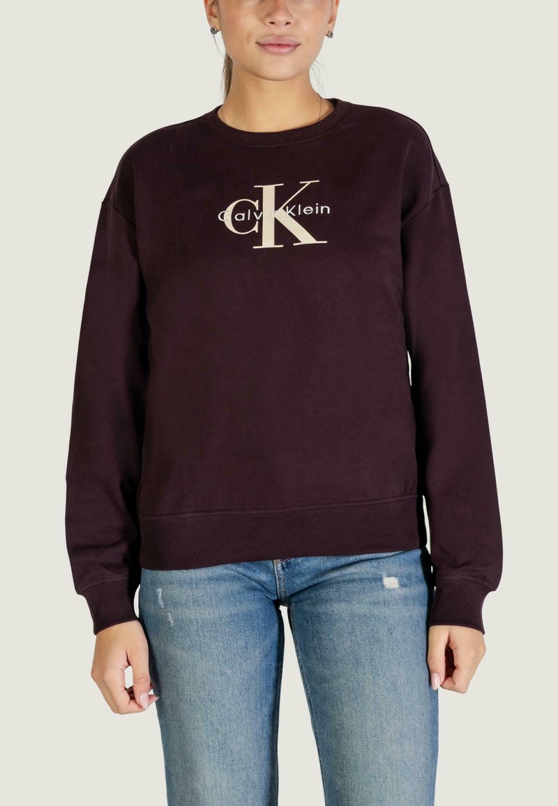 Ciemnofioletowa bluza z kremowym logo Calvin Klein na przodzie, z okrągłym dekoltem i ściągaczami przy mankietach. W zestawieniu z niebieskimi dżinsami.