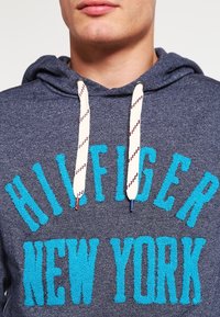 Sudadera con capucha en gris oscuro con el texto "HILFIGER NEW YORK" en textura de toalla azul. Presenta cordones blancos con acentos en rojo y azul.