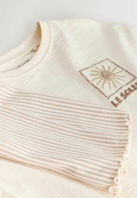 Kremasta majica s potiskom sonca v rjavi barvi. En rokav ima izmenične tanke črte z teksturiranim valovitim robom.