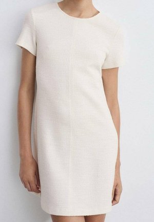 Femme portant une robe crème texturée à manches courtes, longueur genou, sur un fond clair uni.