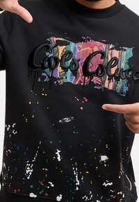 Černá mikina s barevnými vzory postříkání barvou, texturovaným logem a graffiti písmeny s kapkami. Měkký materiál; kulatý výstřih.