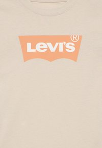 Μπεζ βαμβακερό φανελάκι με μεγάλο πορτοκαλί λογότυπο Levi's και λευκό κείμενο, με μια χαρακτηριστική καμπυλωτή μορφή πάνω από το λογότυπο.