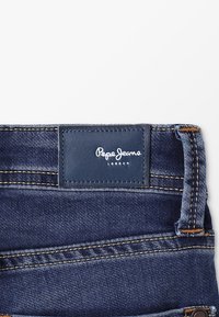 Jeans i mörkblå denim med en texturerad yta. Har en läderlogotyp med texten "Pepe Jeans London" och kontrasterande sömmar.