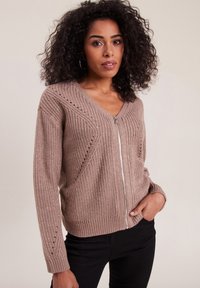 Beige Strickjacke mit V-Ausschnitt und Reißverschluss. Sie verfügt über strukturierte Rippen und offene Maschenverzierungen, lange Ärmel und eine lockere Passform.