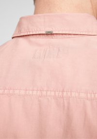 Světle růžová košile s límečkem a prošitým vzorem, s malou kovovou cedulkou na límci a hladkou, měkkou texturou.