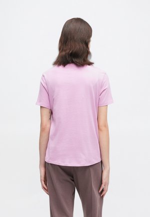 Persoon met schouderlang bruin haar die een lichtroze T-shirt met korte mouwen en een bruine broek draagt, met de rug naar voren tegen een effen witte achtergrond.