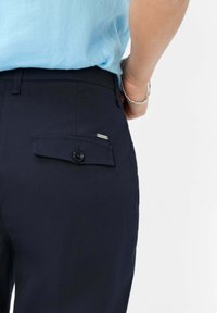 Marineblauwe broek met een achterzak met een flap en knoopsluiting. De stof is glad met een subtiele textuur, gecombineerd met een lichtblauwe top.
