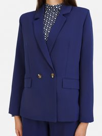 Granatowa dwurzędowa marynarka uszyta z gładkiego materiału, z złotymi guzikami, szerokimi klapami i kieszeniami z przodu. Pod spodem widoczna wzorzysta bluzka.