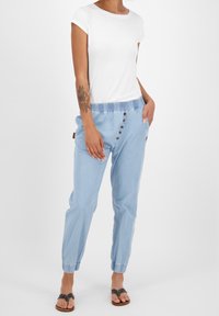 Jasnoniebieskie spodnie na elastycznym pasie z guzikami, w połączeniu z dopasowaną białą koszulką z krótkim rękawem. Modelka nosi płaskie sandały.