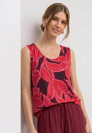 Femme portant un haut sans manches rouge et noir à fleurs avec un pantalon bordeaux, souriante et appuyée contre un mur blanc.