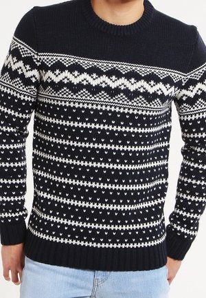 Marineblauer Pullover mit weißem Fair-Isle-Muster, Rundhalsausschnitt, getragen von einer Person in hellblauen Jeans, Hände entspannt an den Seiten.