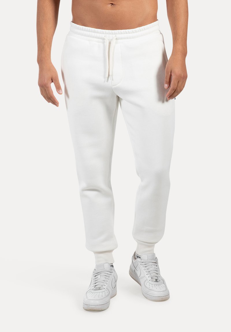 Pantalones de chándal blancos de algodón con puños acanalados, cintura elástica y cordón. Llevados con zapatillas blancas, que presentan un diseño atlético clásico.