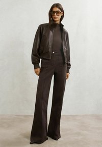 Jachetă bomber din piele maro, helancă închisă la culoare și pantaloni evazați cu talie înaltă. Adaugă o siluetă structurată cu materiale texturate. Ochelari de soare pătrați.