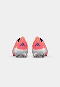 Botas de fútbol Adidas con una parte superior de un coral brillante y suela plateada. Cuenta con una superficie texturizada y tres rayas negras a los lados.