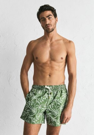Green swim shorts made of lightweight fabric featuring a white tropical leaf pattern. Elastic waistband with drawstring; pockets on the sides.